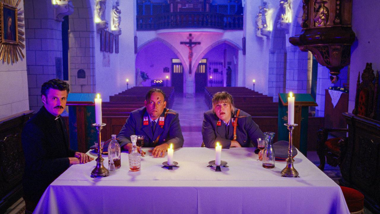 Banyardi, Polizist Hannes und Polizistin Gerti sitzen an einem festlich gedeckten Tisch mit Kerzen in einer Kirche.