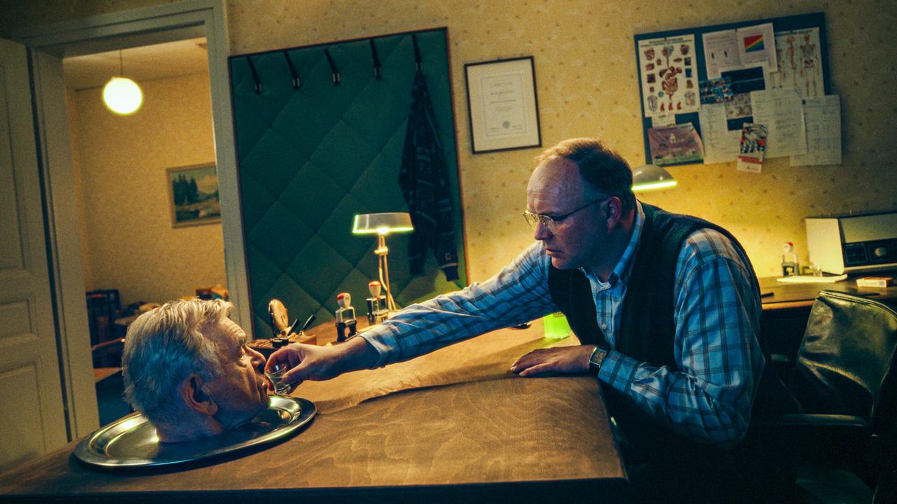 In einem Büro beugt sich Feist Jr. über einen Schreibtisch und hält Vater Feist, dessen Kopf auf einem Silbertablett liegt, ein Glas an den Mund.