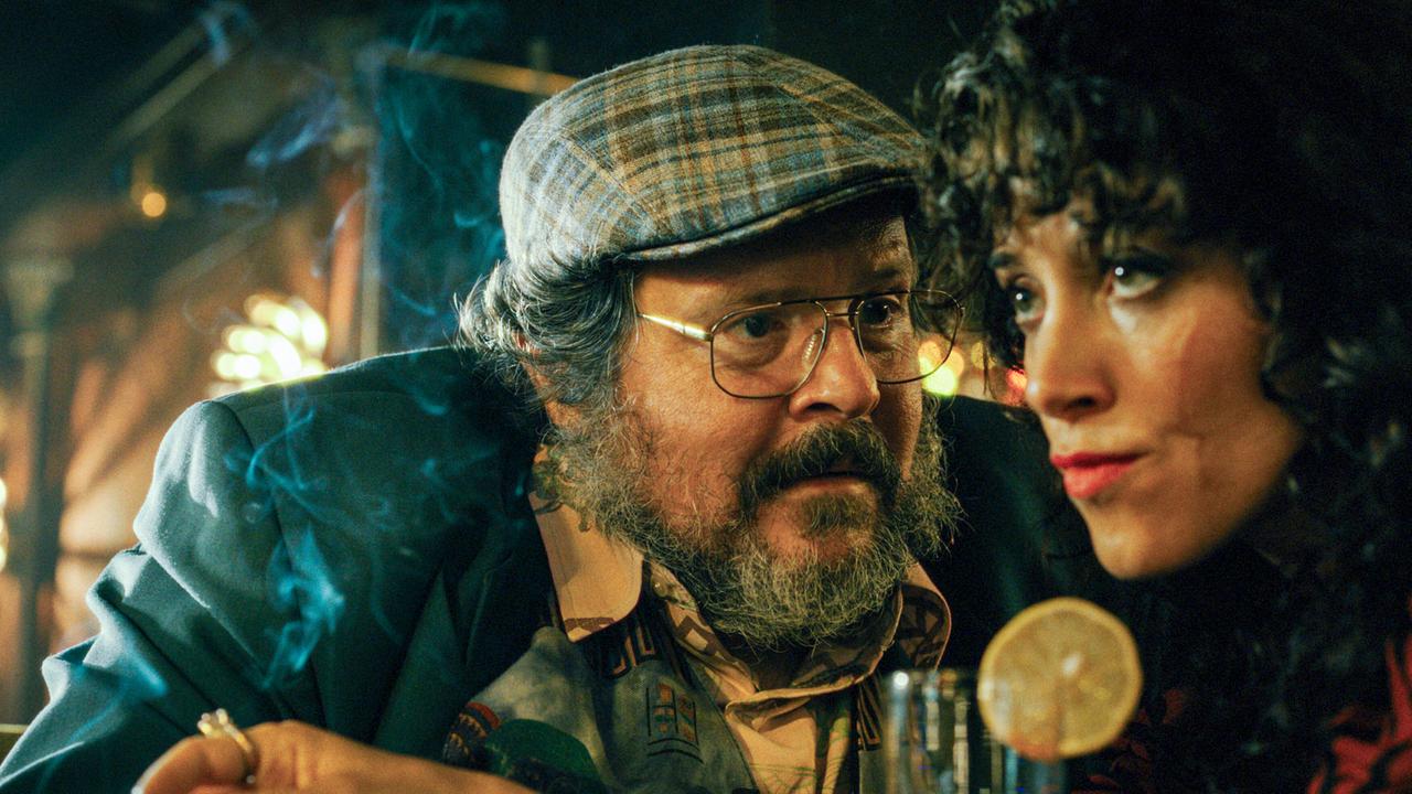 Frosch und Ronda sitzen an der Bar eines Lokals, umgeben von Rauch, Gläsern und gedämpftem Licht.