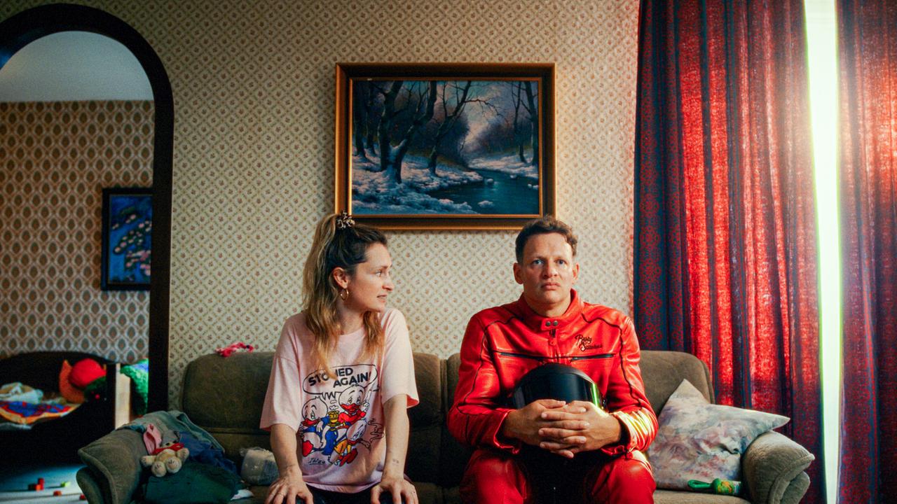  Babs Tschach und Kevin sitzen nebeneinander auf einem Sofa in einem Wohnzimmer mit gemusterter Tapete und Gemälden.