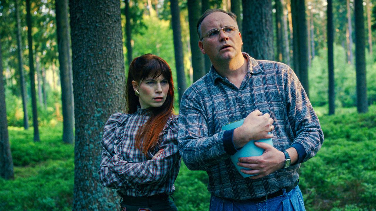 Deborah Feist und Feist Jr., mit einem blauen Behälter in der Hand, stehen im Wald.