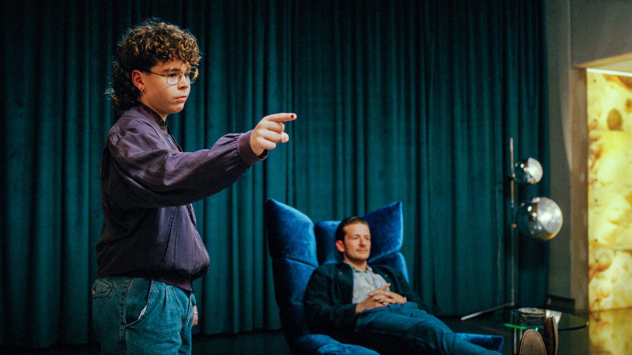 In einem Raum mit dunklen Vorhängen steht der junge B.B. gestikulierend, während ein Mann im Hintergrund entspannt in einem Polstersessel liegt.