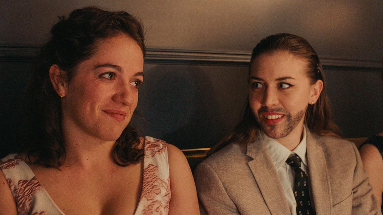 Theresa in einem Kleid und Mira mit aufgemaltem Vollbart und im Anzug sitzen nebeneinander in einem Saal.