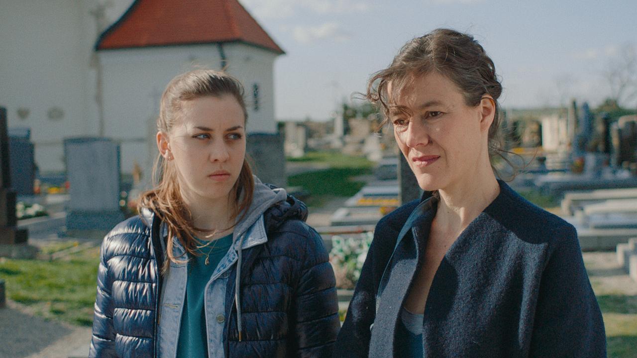 Mira steht mit ihrer Mutter auf einem Friedhof bei sonnigem Wetter.