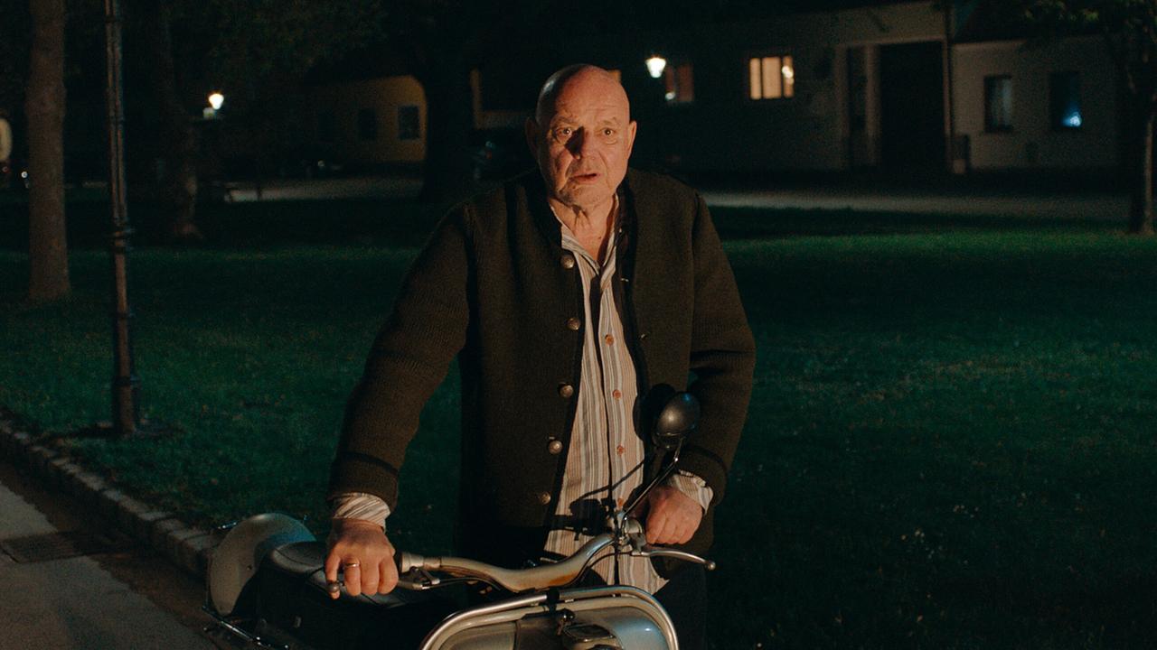 Wolfgang Böck in der Rolle als Opa steht nachts neben einem Motorrad auf einem Weg vor einer Wiese, dahinter eine Häuserreihe.