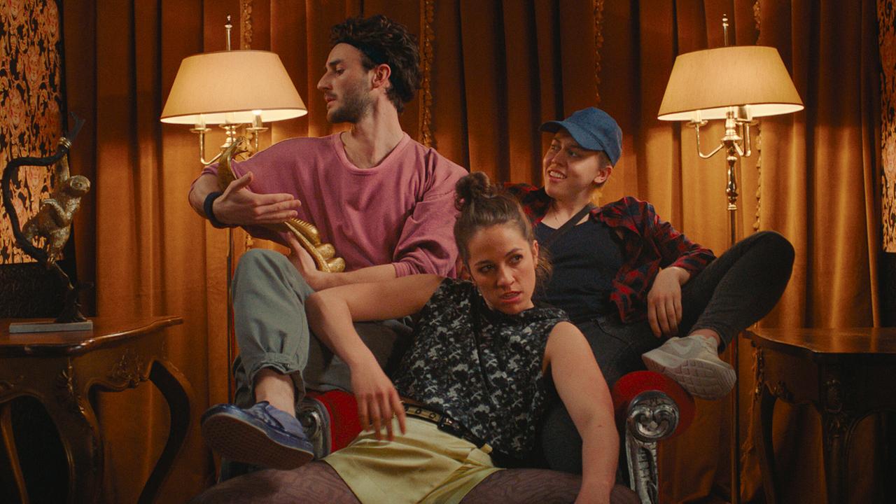 Paul, Theresa und Mira  sitzen zu dritt in einem Coachsessel in einem prunkvoll eingerichteten Raum mit warmem Licht.