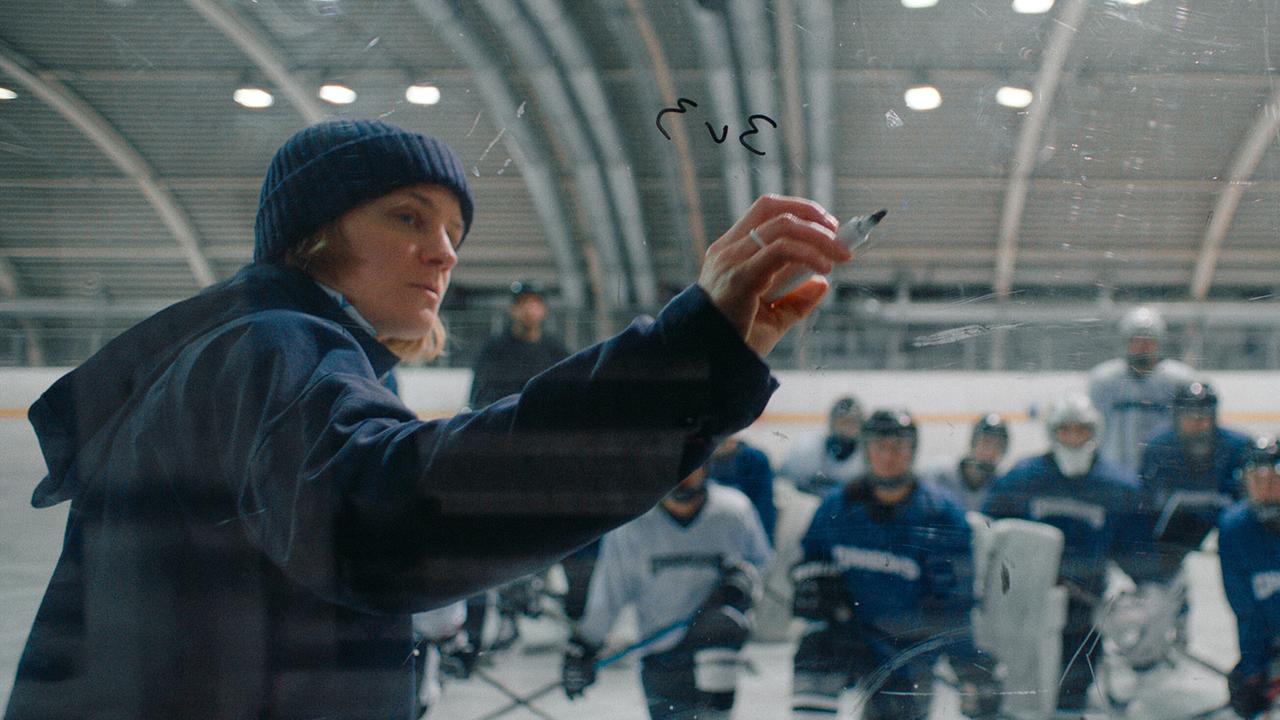 Eine Trainerin zeichnet etwas auf eine Glasscheibe, während ein Eishockeyteam zusieht.