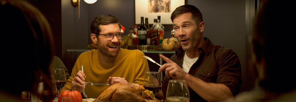 Zwei Männer sitzen bei einem Abendessen nebeneinander und unterhalten sich prächtig mit den anderen Gästen, Bobby schaut Aaron lachend von der Seite an