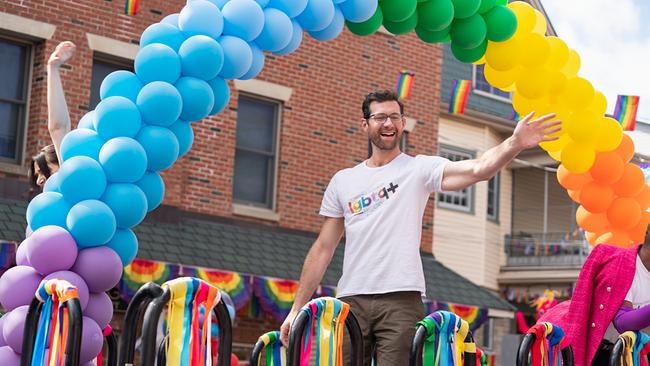 Billy Eichner steht als Bobby auf einem Wagen bei der Regenbogenparade unter bunten Luftballons, fröhlich lächelnd winkt er und trägt ein T-Shirt mit buntem lgbtq+ Aufdruck 