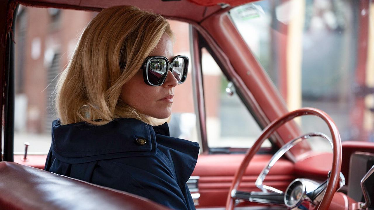 Elizabeth Banks sitzt als Joy - mit schulterlangen blonden Haaren, in einem blauen Mantel mit dunkler Sonnenbrille am Steuer eines geparkten Wagens