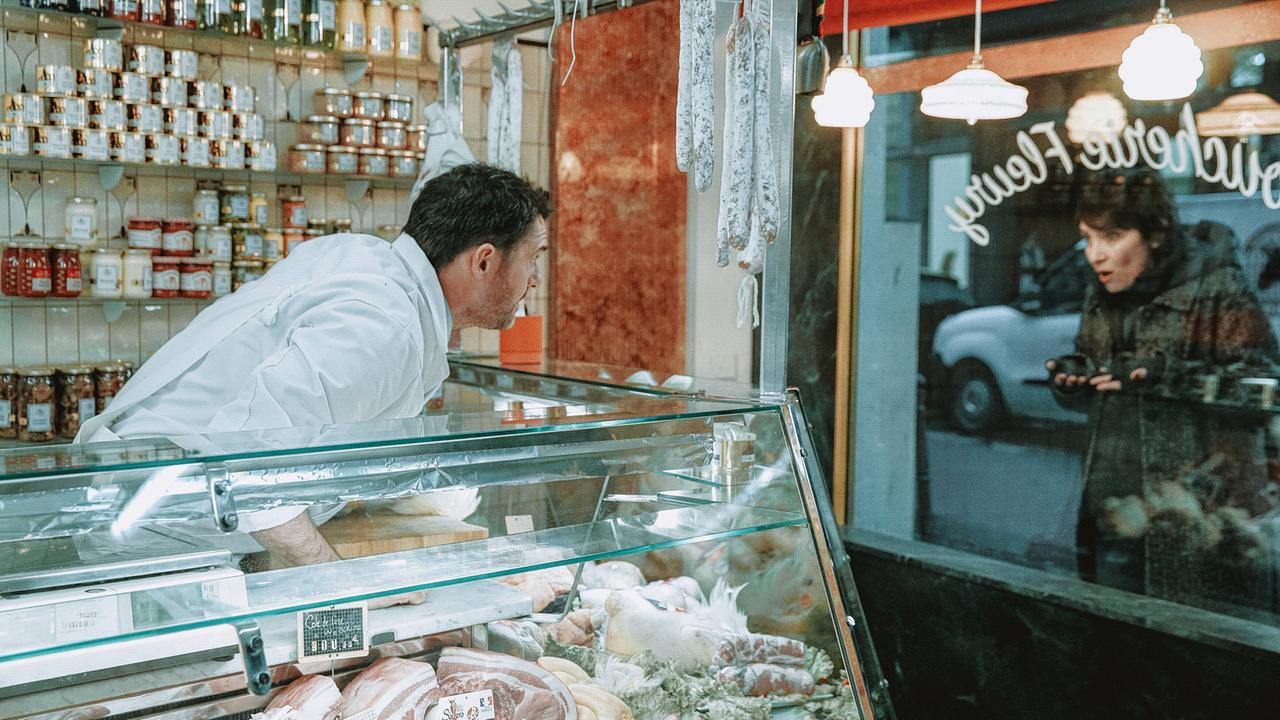 © ORF/Vuelta Entertainment/Les Films du Cap/MM Films/Orange Studio/PIXELLEPHOTO Martial Toussaint in weißer Berufskleidung bückt sich an der Fleischtheke, um durchs Schaufenster mit Charly Fleury Blickkontakt aufnehmen, die vor dem Geschäft steht