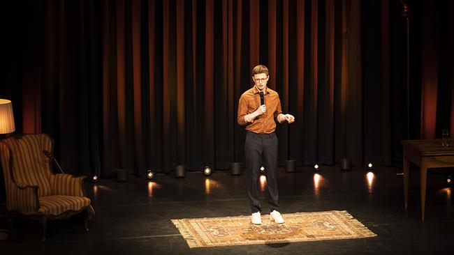 © ORF/Hoanzl/Nikolaus Ostermann Christoph Fritz steht mit einem Mikrofon auf einer wohnzimmerartig eingerichteten Bühne vor einem dunklen Vorhang.