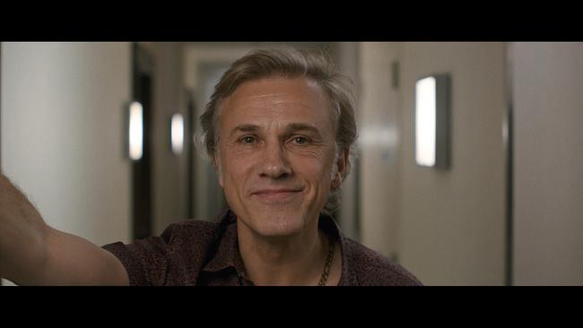 Christoph Waltz steht in einem Hotelgang und macht ein Selfie, lächelnd blickt er freundlich direkt in die Kamera