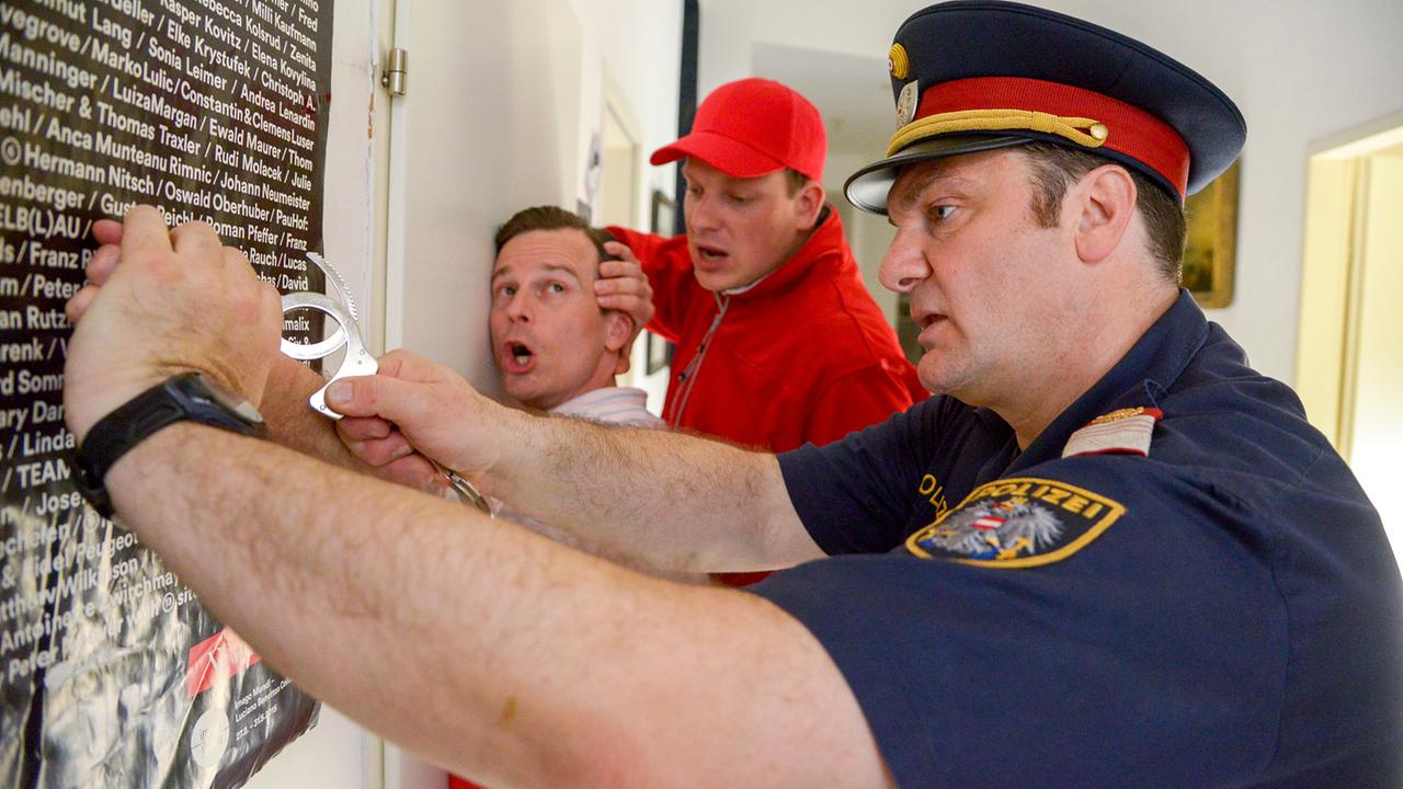 Martin Leutgeb als Polizist legt Mathias Hacker Handschellen an, während Holger Schober in roter Arbeitskleidung danebensteht.