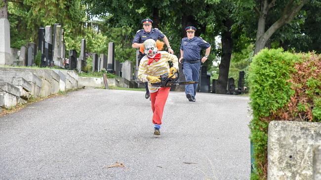 © ORF/Hubert Mican Eine als Clown verkleidete Person rennt mit einer Kettensäge eine Friedhofsallee hinunter, während die zwei Polizisten Roman Mohacsi und Mathias Gerber hinterherlaufen.