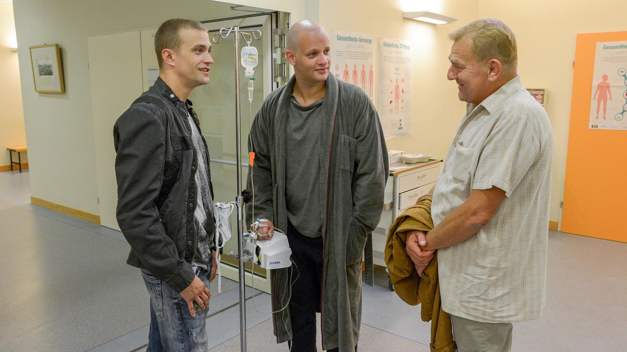 © ORF/Hubert Mican In einem Klinikflur stehen Sylvester Thaler und Wilmer Eberts bei Florian Fehenberger, der an einem Infusionsständer hängt.