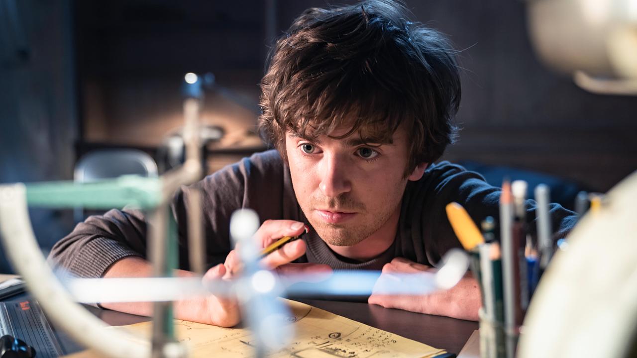 Freddie Highmore als Thom kniet nachdenklich an einem Schreibtisch, er hält einen Stift in der Hand, vor ihm ein Blatt mit Notizen und Formeln