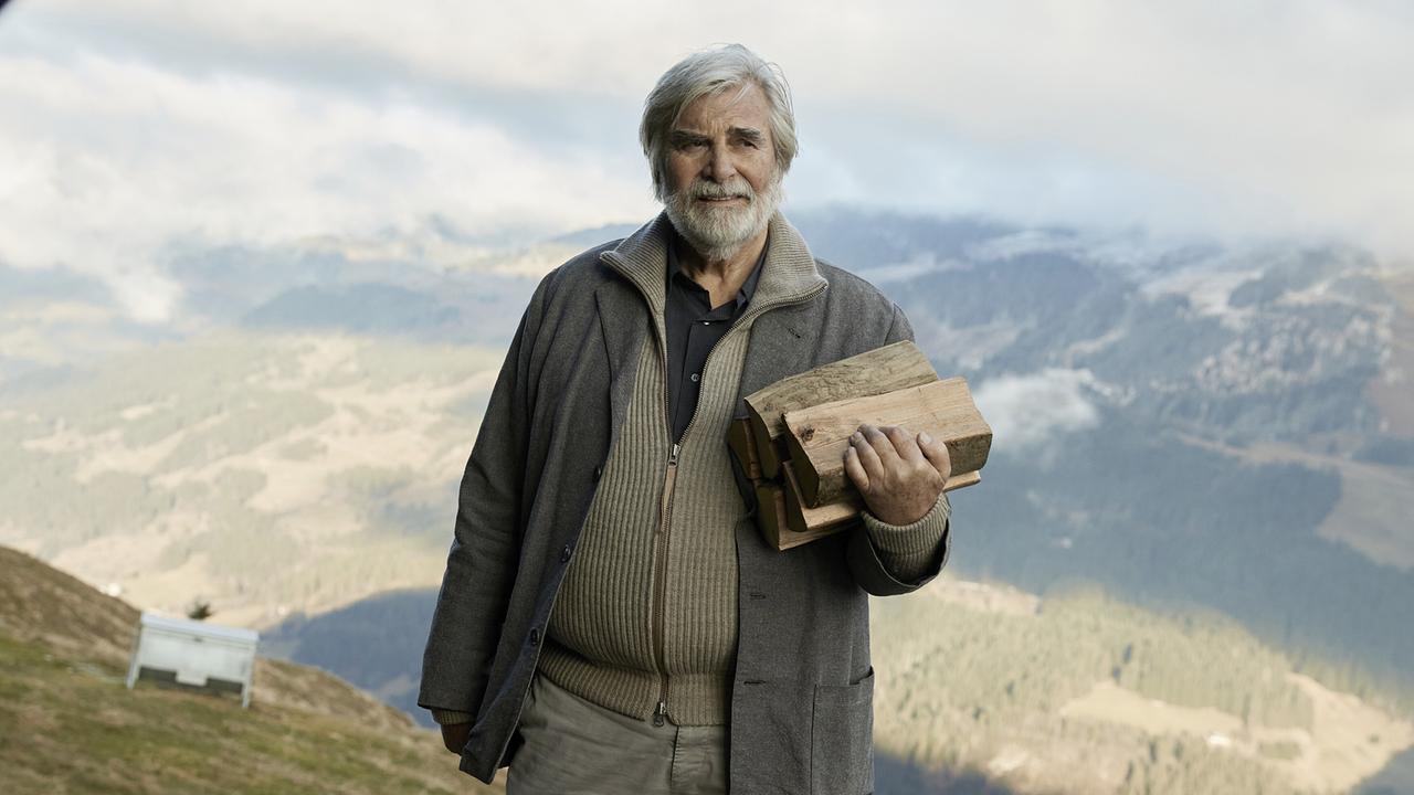 Peter Simonischek als Bernhard auf einer Alm, in Strickjacke und Janker gekleidet hält er Holzscheite im Arm