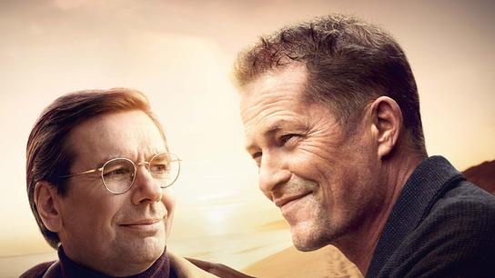 Michael Maertens als Arthur mit Brille und Til Schweiger als Felix stehen nebeneinander, beide lächeln freundlich und blicken aneinander vorbei