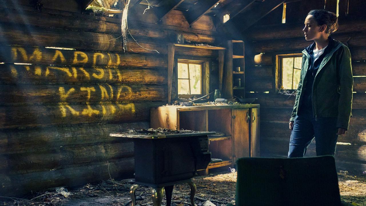 Eine Frau steht in einem von einem Brand zerstörten Raum einer Blockhütte, auf der Wand steht in gelben Blockstaben: Marsh King