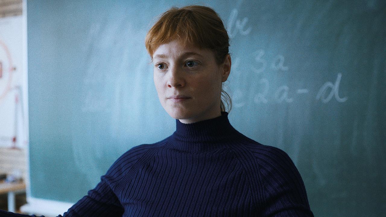 Leonie Benesch steht als Carla Nowak im blauen Rollkragenpullover vor einer Schultafel, auf der mit Kreide mathematische Gleichungen aufgeschrieben sind, gefasst blickt sie in den Klassenraum