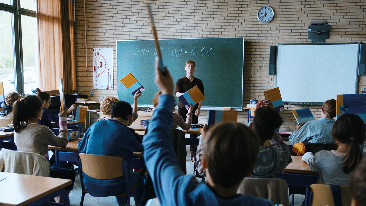 Leonie Benesch als Carla Nowak vor der Tafel im Klassenzimmer, Schülerinnen und Schüler halten ihre Lehrbücher in die Luft