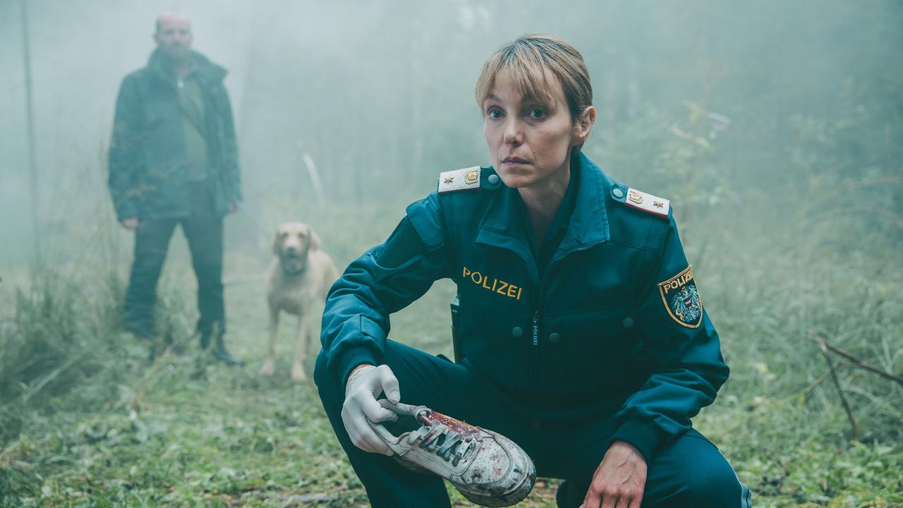 © ORF/ARD Degeto/Mona Film/Victoria Herbig Im Moor: Polizistin Pia Krauss hält einen blutverschmierten Turnschuh in der Hand, der möglicherweise der Vermissten gehört. Hinter ihr steht Gruber mit seinem Hund, der den Schuh gefunden hat.