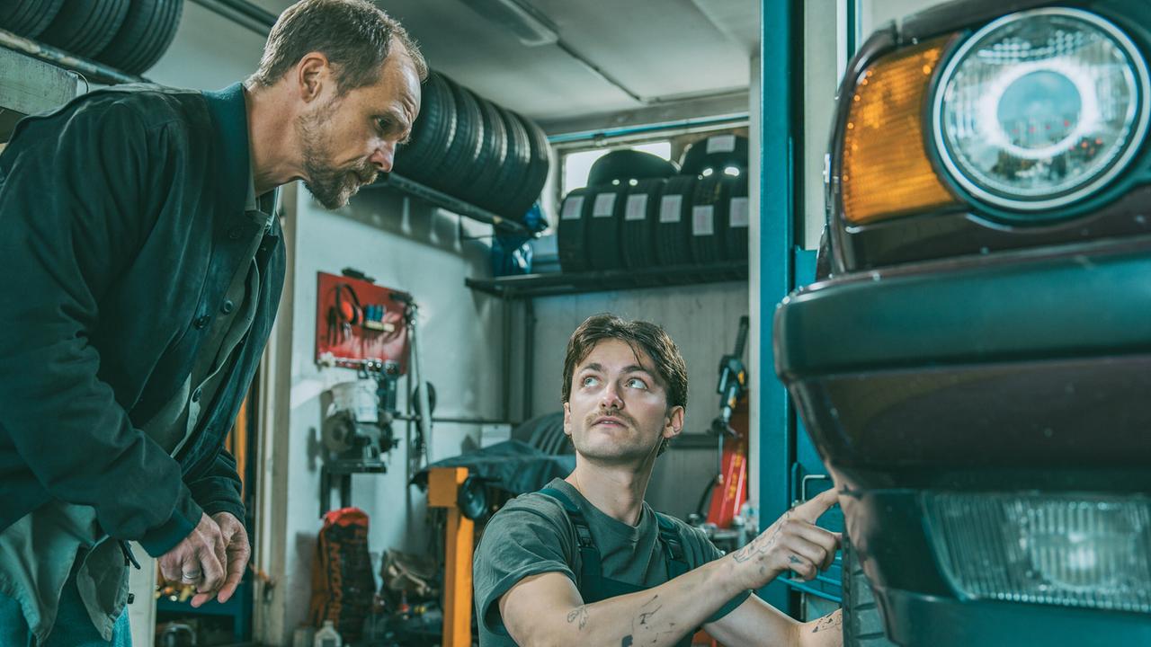 © ORF/ARD Degeto/Mona Film/Victoria Herbig In der Werkstatt von Broszeks Autohandel: Jonas Kraus und Mechaniker Roman unterhalten sich über die Schäden an einem Auto, das Roman gerade repariert.