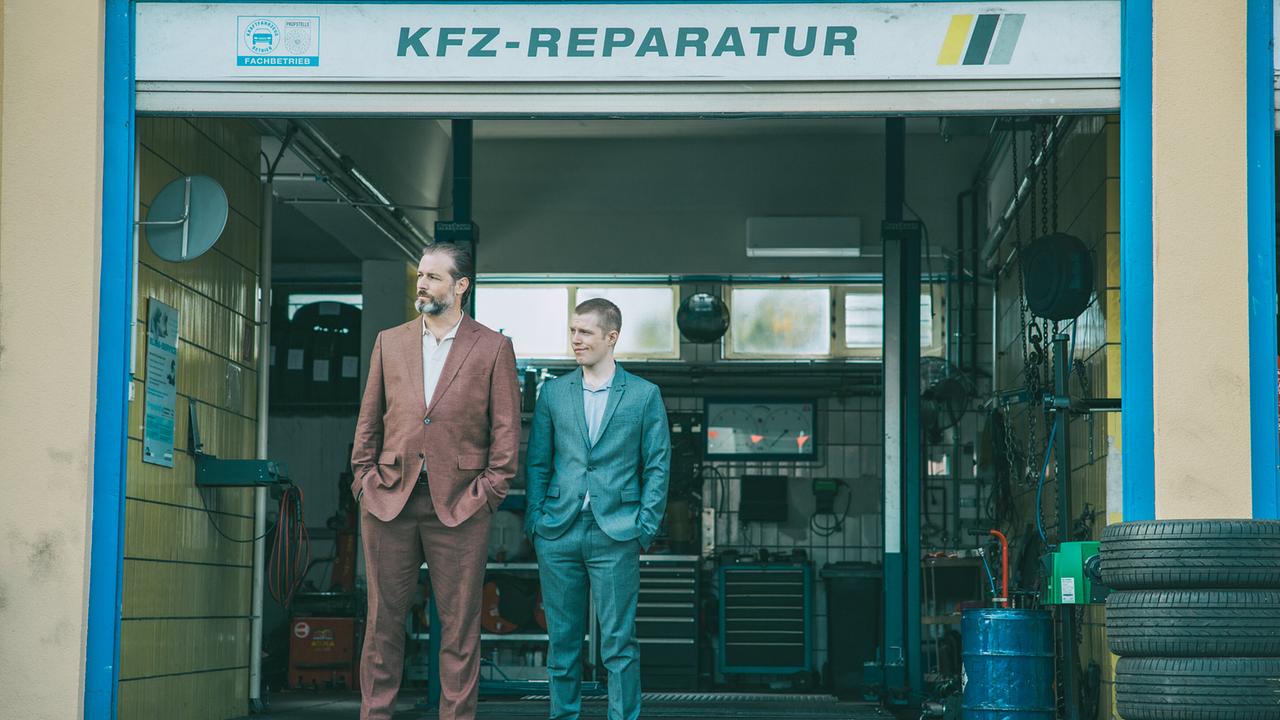 © ORF/ARD Degeto/Mona Film/Victoria Herbig Autohausbetreiber Andreas Broszek und sein Mitarbeiter Felix stehen im Eingang zur Werkstatt und sehen zu, wie Axmann vom Hof fährt.