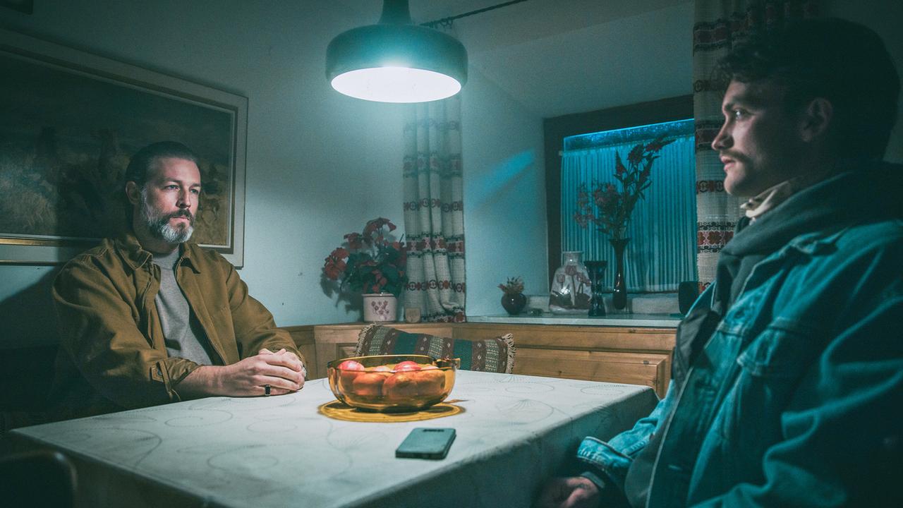 Broszek droht Roman in dessen Küche, während sie gemeinsam am Küchentisch sitzen.