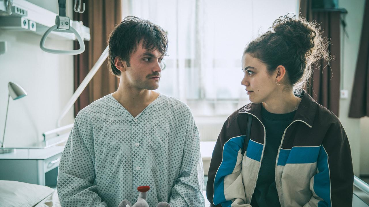 Vivian und Roman sitzen auf seinem Krankenbett. Sie hat ihm ein Stofftier als Geschenk mitgebracht.