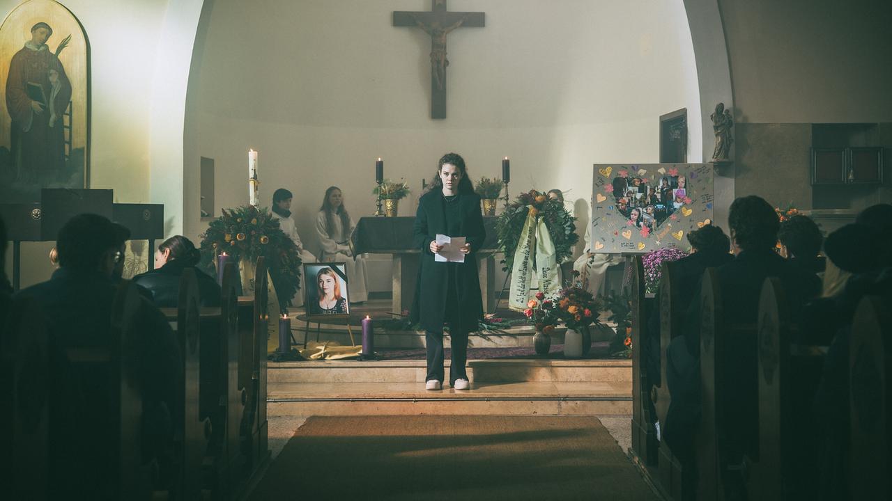 Vivian hält in einer Kirche vor Trauergästen eine Abschiedsrede neben dem Bild der Verstorbenen Cousine Amelie.