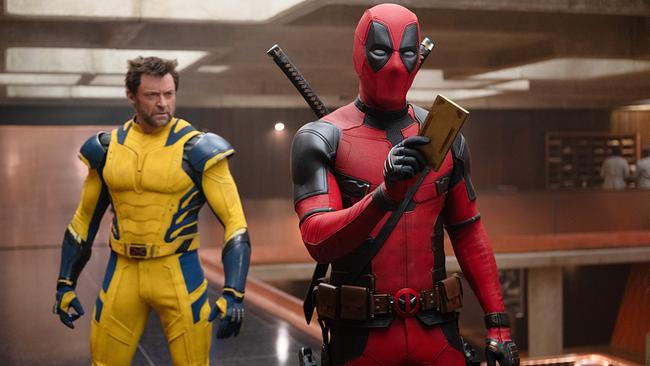 Hugh Jackman als Wolverine in einem gelb-blauen Anzug und Ryan Reynolds als Deadpool in rot-schwarzem Anzug stehen in einem Raum, Deadpool hält etwas in der Hand, das er seinem Gegenüber zeigt