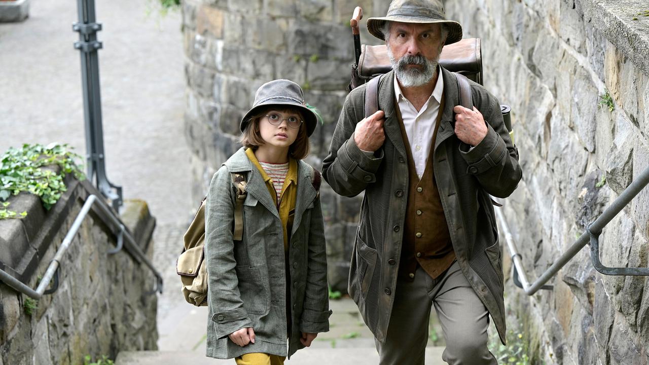 Christoph Maria Herbst als Buchspazierer Carl Kolhoff mit ledernem Rucksack steigt er in Begleitung von Yuna Bennett als Schascha eine steinerne Treppe hinauf