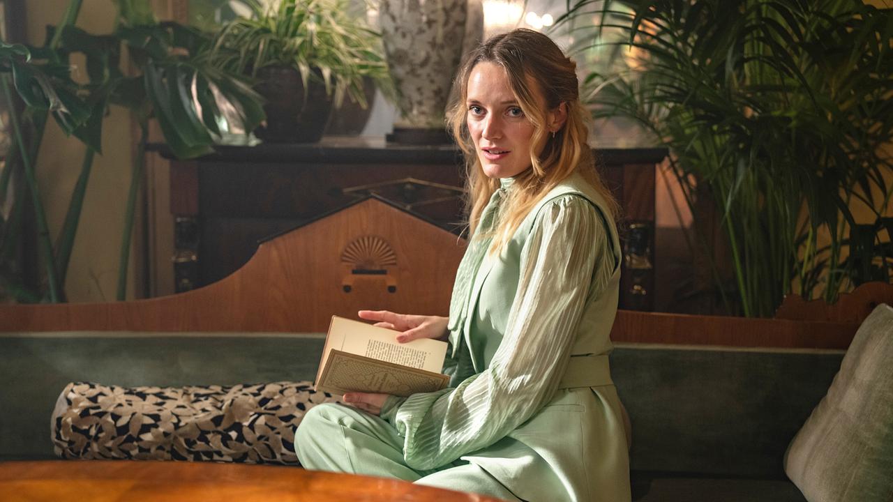 Hanna Hilsdorf als Effi in einem hellgrünen Kleid auf einer Bank in einem Wohnzimmer mit einem Buch sitzt, ihr Platz ist umgeben von Zimmerpflanzen