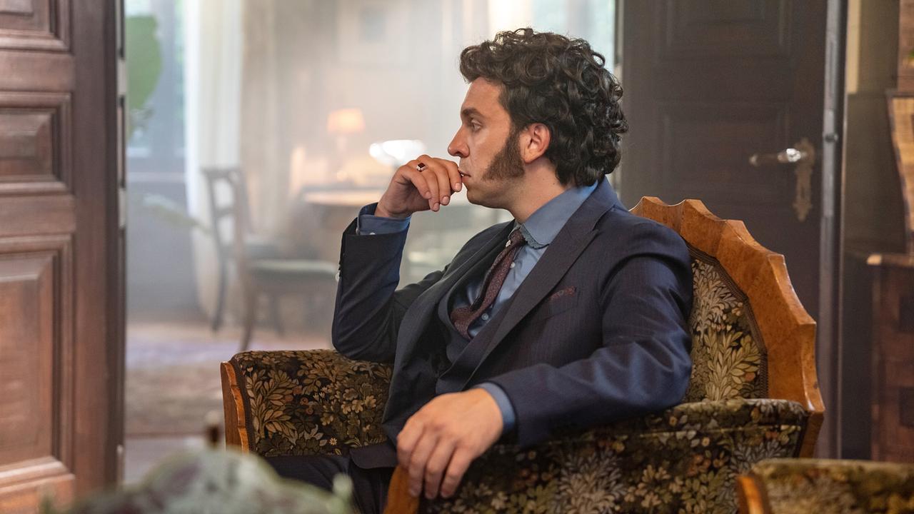 Edin Hasanovic als Mister Darcy mit lockig gewelltem Haar und Backenbart, in einem stilvoll eingerichteten Wohnzimmer sitzt er nachdenklich in einem Lehnsessel