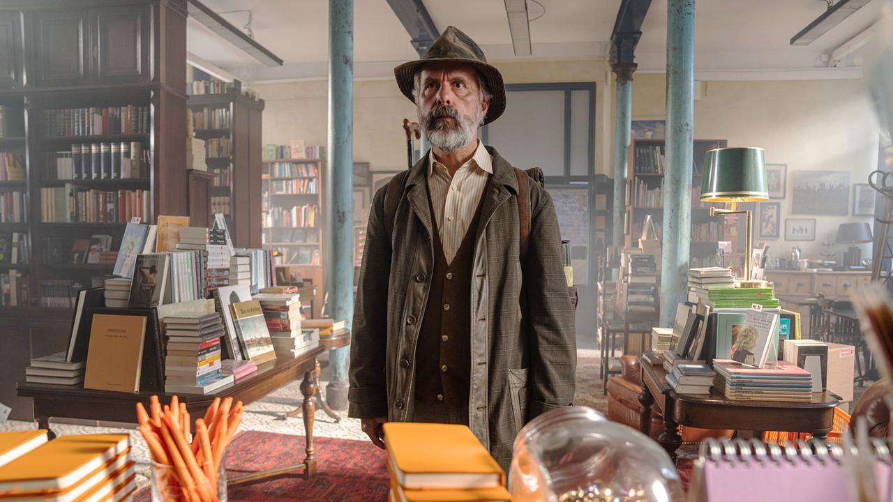 Christoph Maria Herbst als Carl Kolhoff in einer alten Buchhandlung, altmodisch mit Hemd, Weste, Jacke und Hut bekleidet, steht er mit seinem Rucksack vor der dem Kassatisch