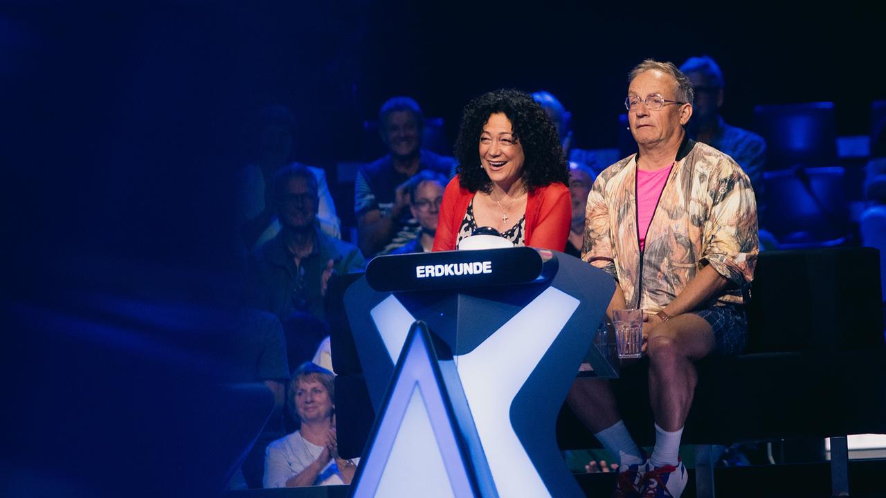 © ORF/ZDF/Thomas Kierok Barbara Wussow und Wigald Boning sitzen am blau beleuchteten Ratetisch der Kategorie „Erdkunde“ vor dem Studiopublikum bei "Der Quiz Champion".