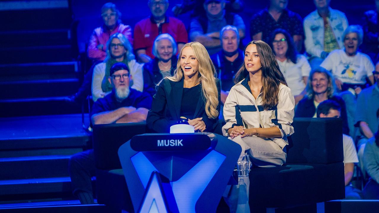 © ORF/ZDF/Thomas Kierok Melissa Naschenweng und Vanessa Mai sitzen am blau beleuchteten Ratetisch der Kategorie „Musik“ vor dem Studiopublikum bei "Der Quiz Champion".