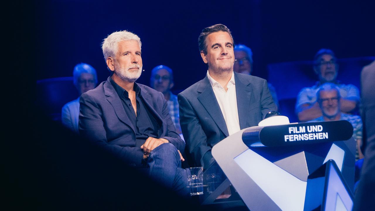 © ORF/ZDF/Thomas Kierok Dirk Stermann und Bastian Pastewka sitzen am blau beleuchteten Ratetisch der Kategorie „Film und Fernsehen“ vor dem Studiopublikum bei "Der Quiz Champion".