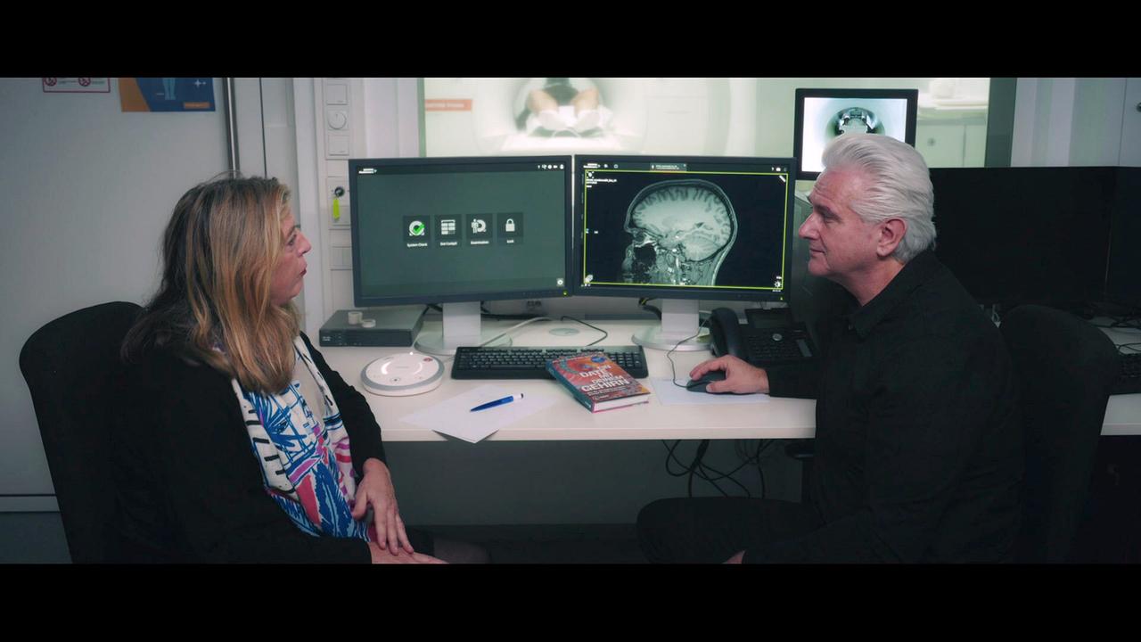 Prof. Anna Buchheim ( Universität Innsbruck) im Gespräch mit Dr.Dr. Damir del Monte an einem Schreibtisch vor einem Computer, der medizinische Gehirnaufnahmen zeigt, dahinter ein Untersuchungsraum.