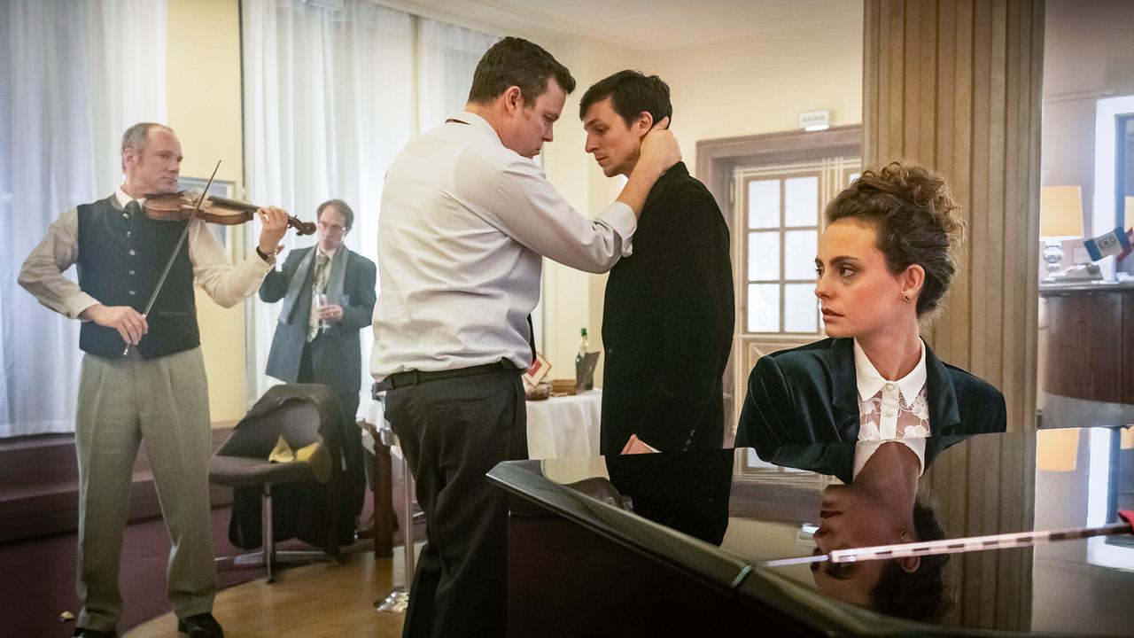In einem Saal spielt ein Geiger, während im Vordergrund der junge Walter Lobrecht den jungen Kurt Lehmann am Kragen packt und eine Frau am Klavier sitzt.