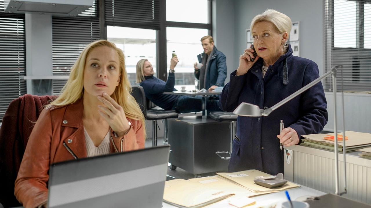 Im modernen Kommissariatsbüro telefoniert Henriette Wolf neben Akten und Schreibtischlampe, während Penny Lanz am Laptop sitzt und im Hintergrund Carl Ribarski und Simon Steininger zu sehen sind.
