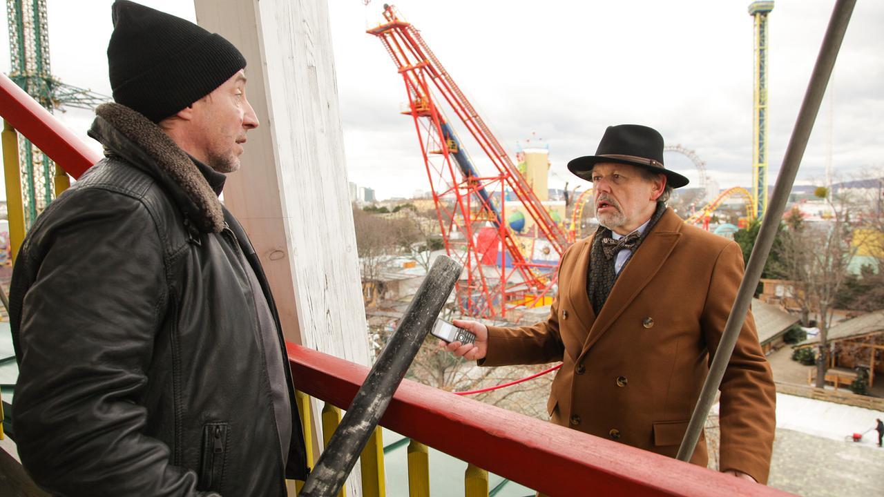 Auf einer erhöhten Plattform in einem Vergnügungspark steht Michael Havranek mit einem Handy in der Hand vor Kurt Lehrmann, der einen Schlagstock in der Hand hat.
