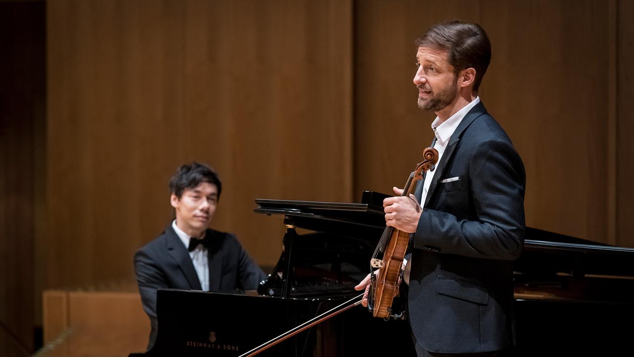 Auf einer Bühne steht der Geiger Philipp Baumgarten mit Instrument vor einem Flügel, an dem der Pianist Karsten sitzt.