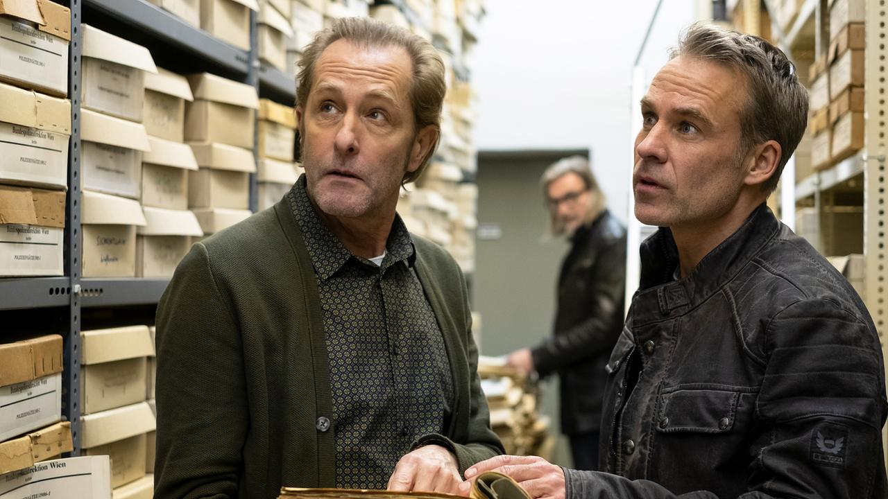 Franz Wohlfahrt und Jan Maybach stehen in einem Archivgang zwischen Unterlagen, während im Hintergrund Carl Ribarski Akten durchblättert.