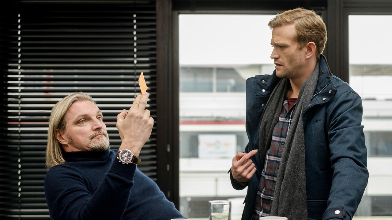 Im Büro zeigt der sitzende Carl Ribarski ein kleines Stück Papier, während Simon Steininger daneben am Tisch steht; vor ihnen stehen Tassen und ein Wasserglas.