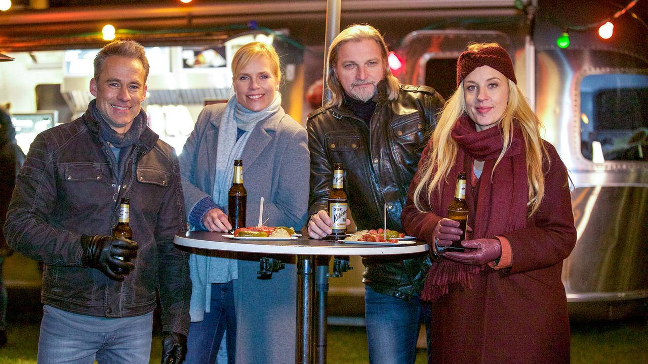 Jan Maybach, Ina Zimmermann, Carl Ribarski und Penny Lanz stehen abends draußen an einem Stehtisch mit Bierflaschen und Würsteln vor einem Foodtruck.