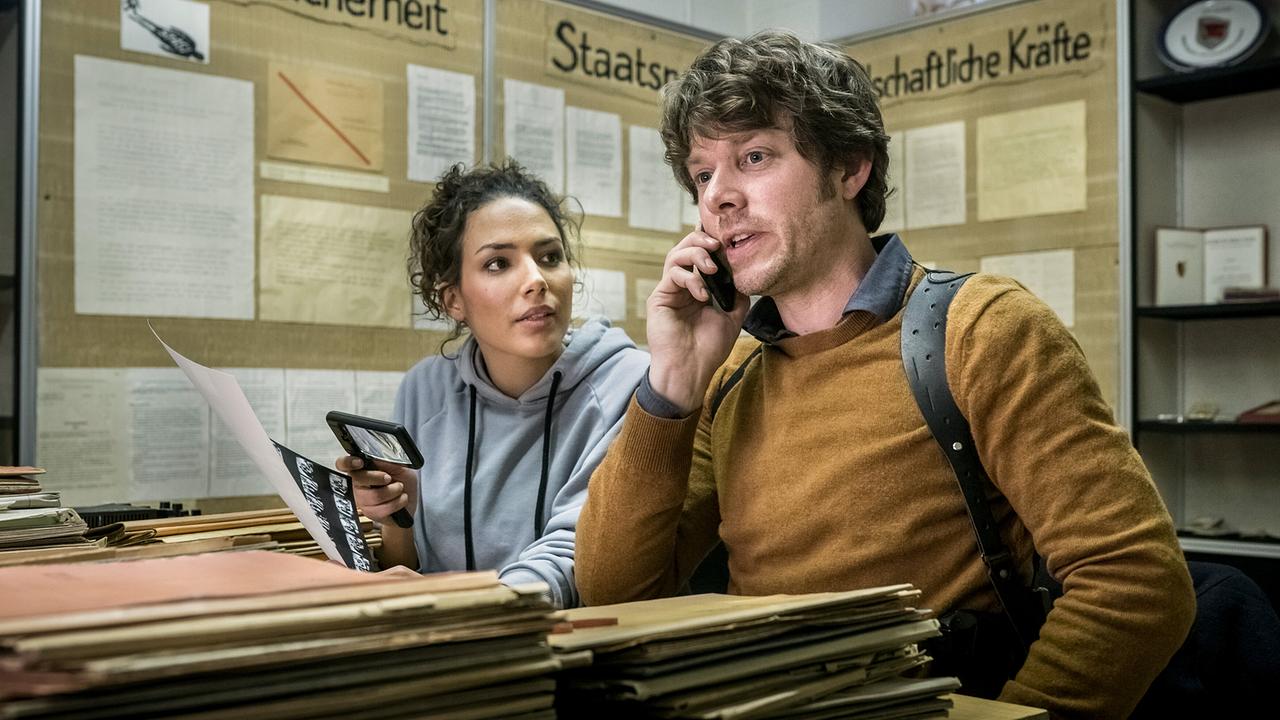 Zwischen hohen Aktenstapeln telefoniert Tom Kowalski, während Kim Nowak neben ihm ein Dokument in der Hand hält.