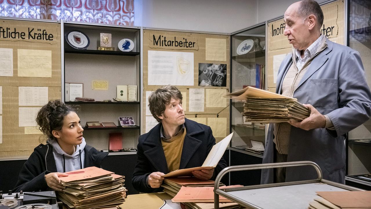 In einem Archivraum mit Vitrinen und vielen Akten bringt ein Archivar weitere Mappen, während Kim Nowak und Tom Kowalski Unterlagen sichten.